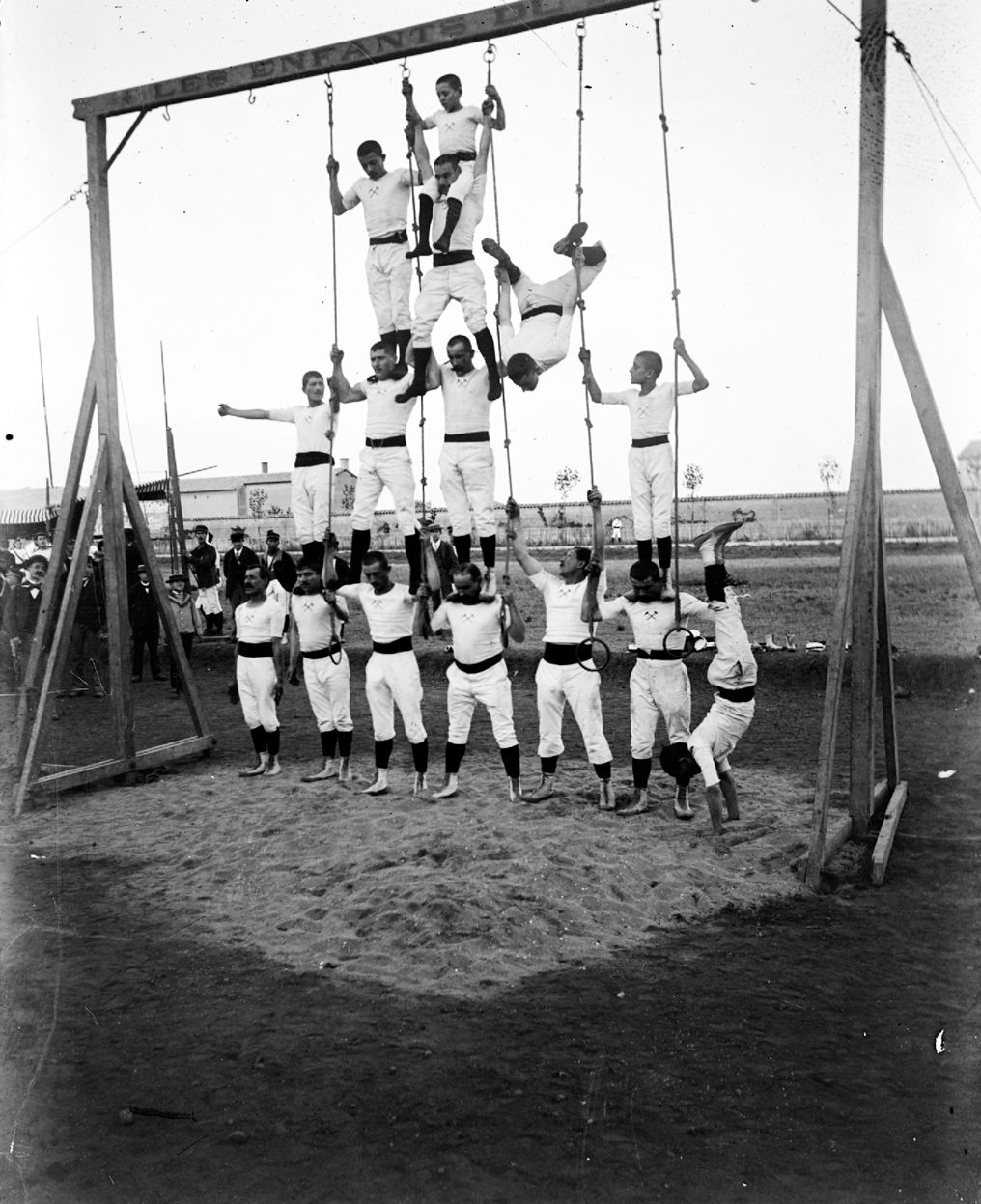 Revue militaire au Grand-Camp (Sathonay-Camp, dans le Rhône) - Gymnastes formant une pyramide humaine sous un portique : photographie négative NB sur verre (vers 1900, cote 10PH/120)