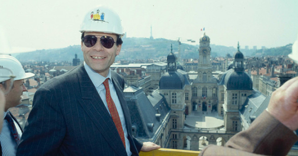 Le Maire Michel noir avec un casque de chantier devant une vue de la Mairie de Lyon